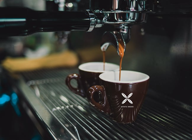 Close-up of two rich espresso shots being brewed into dark brown Xstatic Brews branded ceramic cups from a professional espresso machine. Enjoy the premium coffee experience.
